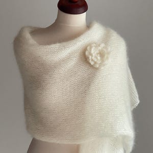 May include: A cream-colored, fluffy shawl draped over a mannequin. The shawl is adorned with a matching flower pin. The fabric appears soft and delicate, with a slight sheen. The overall style is elegant and suitable for special occasions.