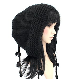 May include: A black knitted hood with long ear flaps and dangling decorative elements. The hood is displayed on a mannequin head, showcasing its shape and texture. The hood's design includes a textured knit pattern and decorative tassels.