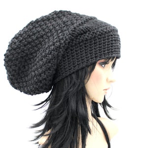 May include: A dark gray, slouchy crochet hat. The hat is made of a thick yarn and has a textured appearance. The hat is shown on a mannequin head with long, black hair. The hat is a stylish accessory for cold weather.