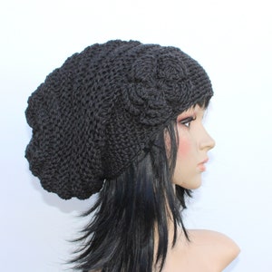 May include: A black crochet hat with a flower design on the side. The hat is made of a thick yarn and has a slouchy fit. The hat is shown on a mannequin head with long black hair.