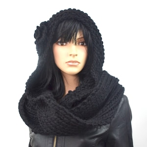 Puede incluir: Un conjunto de capucha y bufanda de punto negro. La capucha presenta una flor decorativa en el lateral y un punto grueso y texturizado. La bufanda es un punto grueso negro a juego, diseñado para llevar alrededor del cuello. El conjunto se muestra en un maniquí.