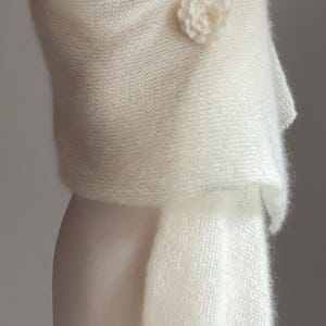 May include: A soft, white, knitted shawl draped over a mannequin. A delicate, white flower pin adorns the shoulder. The shawl has a fluffy texture and a flowing design, perfect for special occasions.