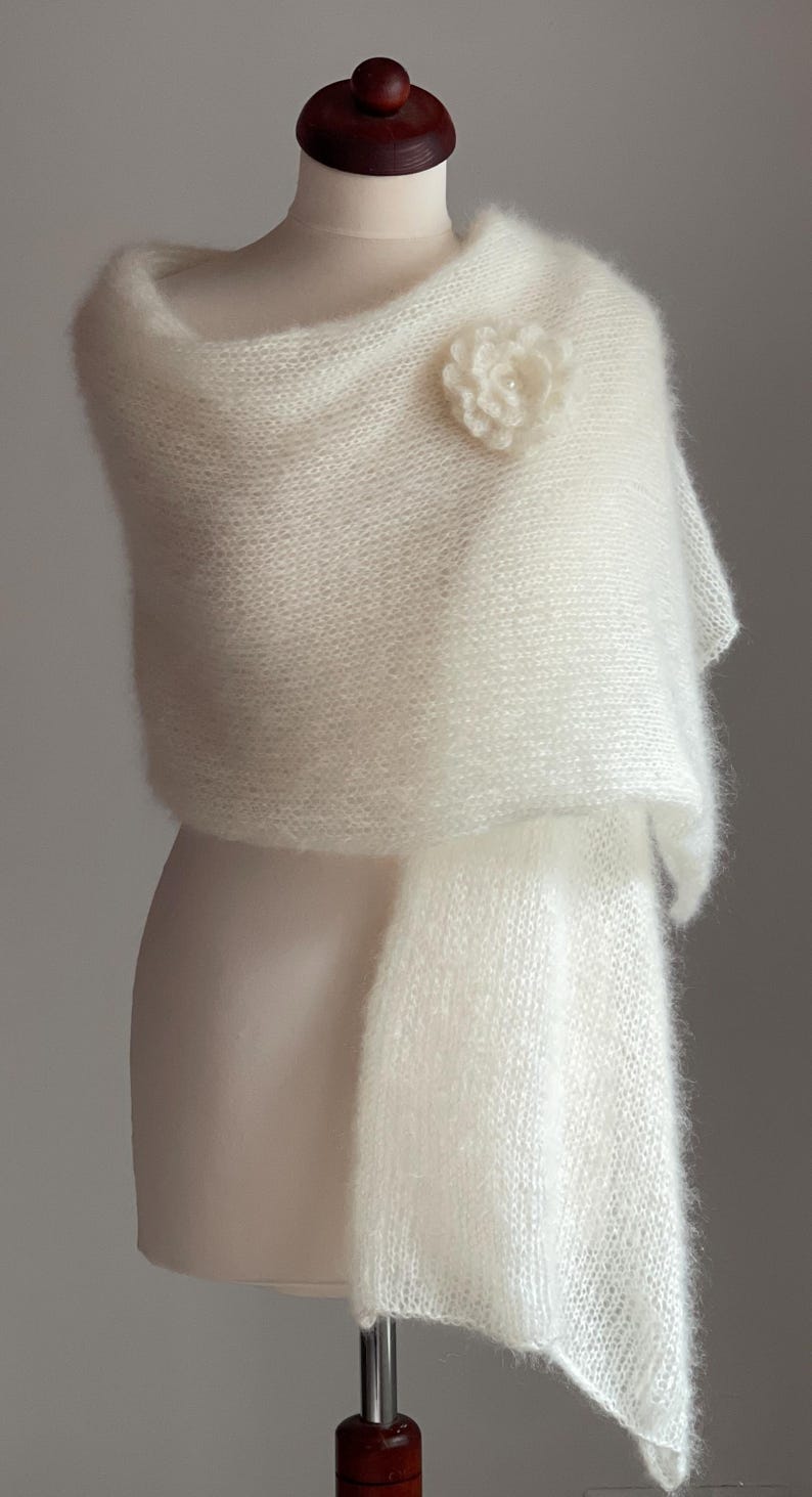 May include: A soft, white, knitted shawl draped over a mannequin. A delicate, white flower pin adorns the shoulder. The shawl has a fluffy texture and a flowing design, perfect for special occasions.
