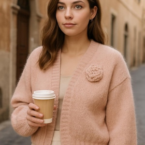 Op de afbeelding: Een vrouw draagt een lichtroze vest met bloemdetail en houdt een papieren koffiebeker vast. Het vest wordt gecombineerd met een crèmekleurige broek. De achtergrond is een Europese straat.