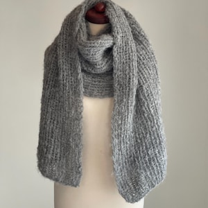 May include: A gray, chunky-knit scarf draped over a mannequin. The scarf is made of a soft, textured yarn and has a loose weave. The scarf is a solid gray color and appears warm and cozy.
