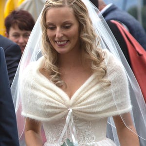 May include: A bride in a white wedding dress with a lace bodice and a cream-colored knitted shawl. She wears a sheer veil and holds a bouquet of pink and white flowers. Her blonde hair is styled with a braid.