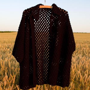 May include: A black, open-weave crochet cardigan hangs on a wooden hanger. The cardigan has a collar and short sleeves. The background is a field of wheat, with a clear sky above.