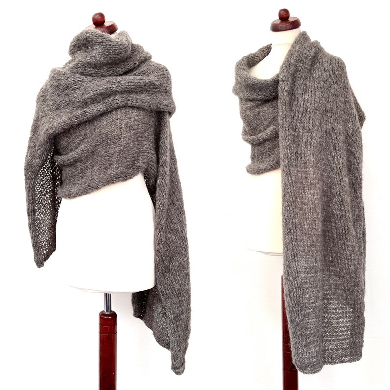 May include: A gray knitted shawl displayed on a mannequin. The shawl is draped to show its texture and shape, with a loose, open weave on one edge. The fabric appears soft and warm, suitable for cooler weather.