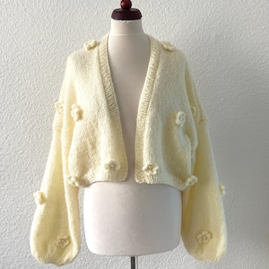 May include: A cream-coloured, open-front cardigan with long, puffy sleeves. The sweater is decorated with small, three-dimensional flower appliqués. The garment is displayed on a mannequin against a plain white background.