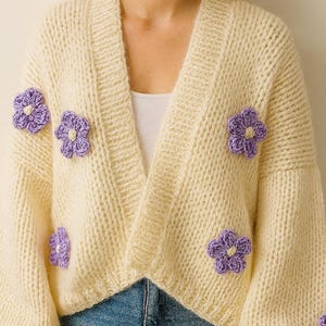 May include: A cream-coloured, chunky knit cardigan decorated with several purple crocheted flowers. The open-front sweater has a V-neck and long sleeves. The model is wearing blue jeans.