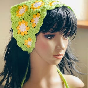 May include: A handmade, green crochet headband featuring a floral pattern with yellow and white accents. The headband is designed with a scalloped edge and ties at the back. The accessory is shown on a mannequin.