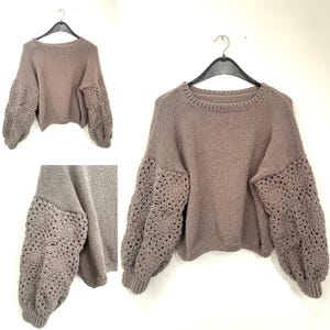 May include: A taupe-coloured jumper with a round neckline and long sleeves. The sleeves feature a crocheted floral pattern, adding a unique texture to the design. The jumper is displayed on a black hanger against a white background.