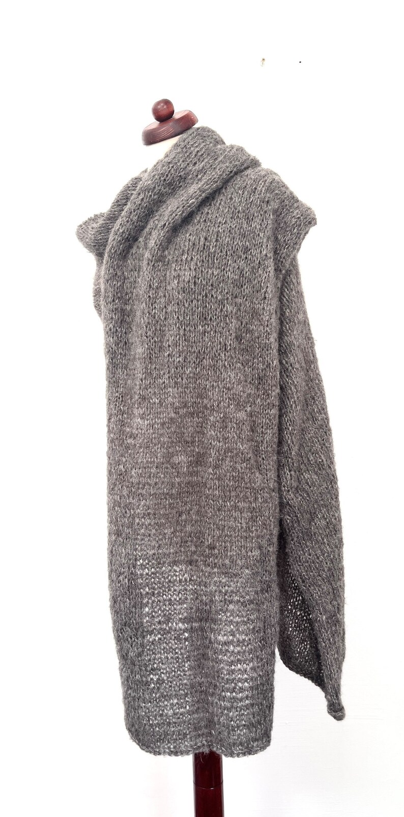 May include: A gray, oversized knitted shawl displayed on a mannequin. The shawl is made of a thick, textured yarn and features a draped design. The color is a muted gray, and the garment appears warm and cozy.