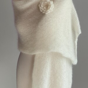 May include: A cream-colored, knitted shawl draped over a mannequin. A white flower pin is attached to the shawl. The shawl is made of a soft, textured material. The mannequin has a wooden head.
