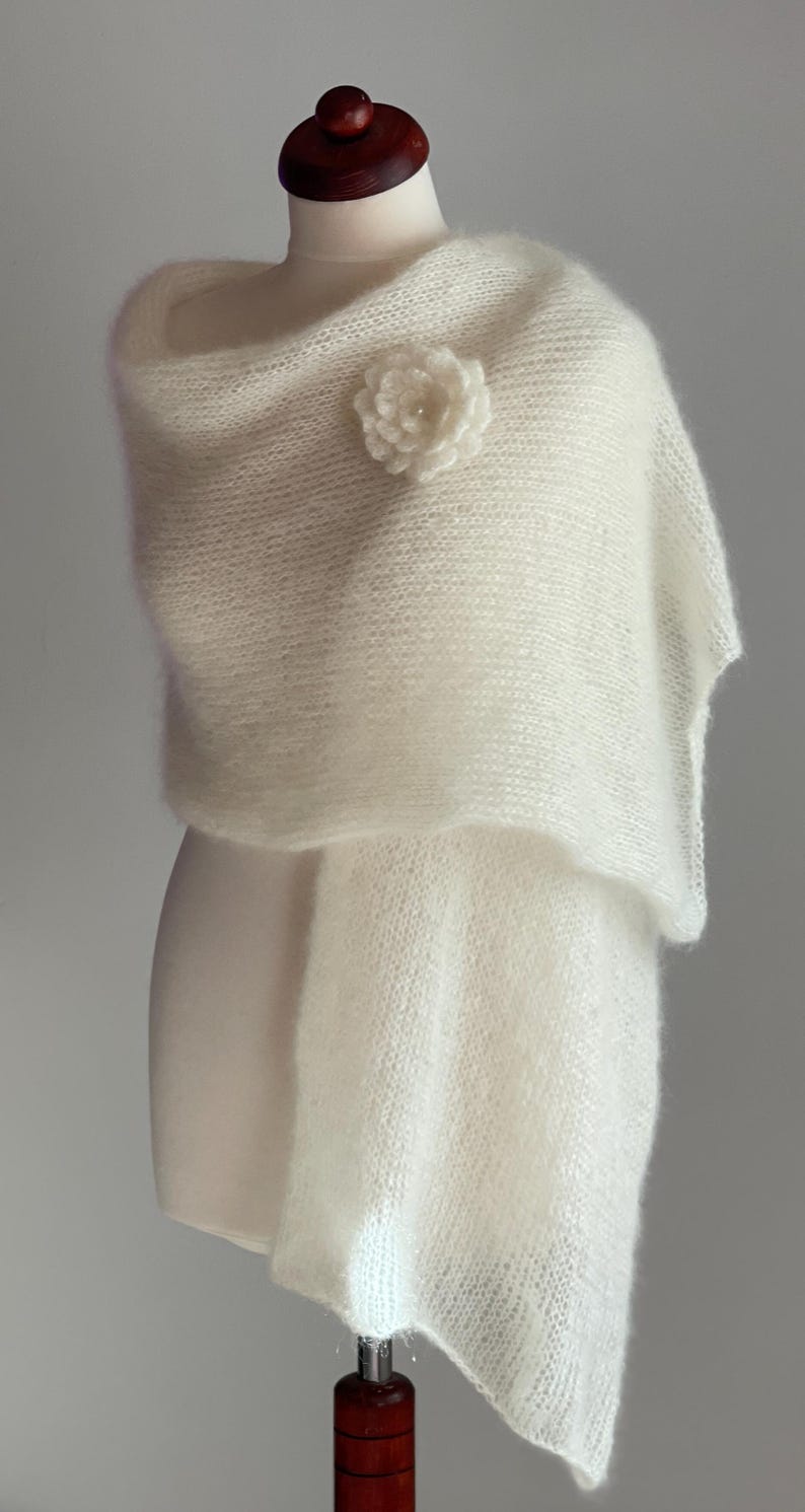 May include: A cream-colored, knitted shawl draped over a mannequin. A white flower pin is attached to the shawl. The shawl is made of a soft, textured material. The mannequin has a wooden head.