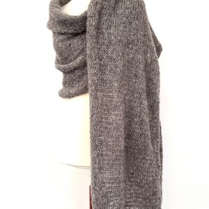 May include: A gray knitted scarf draped over a mannequin. The scarf is made of a thick, textured yarn and has a long, rectangular shape. The scarf is a neutral color, suitable for various outfits. The scarf is displayed against a white background.