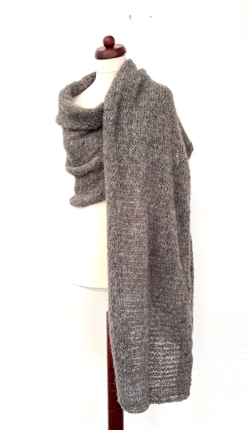 May include: A gray knitted scarf draped over a mannequin. The scarf is made of a thick, textured yarn and has a long, rectangular shape. The scarf is a neutral color, suitable for various outfits. The scarf is displayed against a white background.