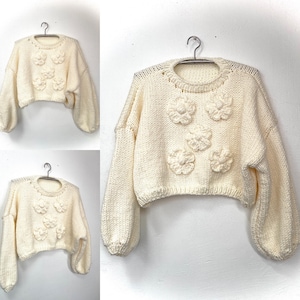 May include: Cream-colored, cropped sweater with a round neckline and long, voluminous sleeves. The sweater features five 3D flower appliqués across the front. The garment is displayed on a hanger against a white background.