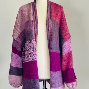 May include: A hand-knitted, open-front cardigan in shades of purple and pink. The sweater features a patchwork design with varying textures and colour blocks. The sleeves are long and the overall style is relaxed and cosy.