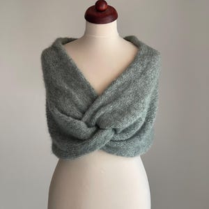May include: A sage green, knitted shawl is draped over a mannequin. The shawl is a soft, fuzzy material and has a twisted knot design in the front. The mannequin has a wooden head and is set against a neutral background.