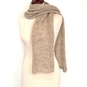 May include: A beige knitted scarf draped over a mannequin. The scarf is a solid color with a textured appearance. The mannequin is a neutral color, and the background is white. The scarf appears to be made of a soft, warm material.