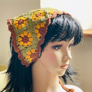 May include: A handmade crochet headscarf in a triangular shape. The head covering features a floral pattern with yellow, brown, and olive green colors. The edges are scalloped and trimmed in brown. The headscarf is displayed on a mannequin.