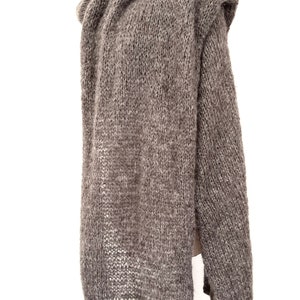 May include: A long, gray knitted hooded sweater displayed on a mannequin. The sweater is made of a thick, textured yarn and has long sleeves. The hood is draped over the mannequin's shoulders. The sweater is a neutral color, suitable for various outfits.
