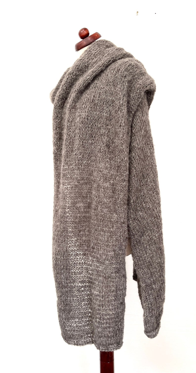 May include: A long, gray knitted hooded sweater displayed on a mannequin. The sweater is made of a thick, textured yarn and has long sleeves. The hood is draped over the mannequin's shoulders. The sweater is a neutral color, suitable for various outfits.