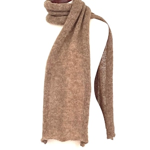 May include: A light brown knitted scarf displayed on a white mannequin. The scarf is draped over the mannequin's shoulders and hangs down. The scarf has a simple, textured knit pattern and a fringed edge. The scarf is made of a soft, warm material.