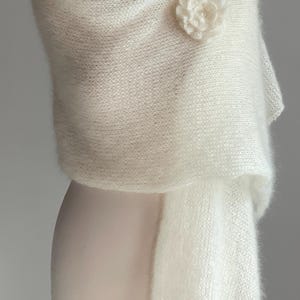 May include: A white, fluffy shawl draped over a mannequin. The shawl is secured with a flower-shaped brooch. The fabric appears soft and delicate, with a slight sheen. The overall aesthetic is elegant and suitable for special occasions.