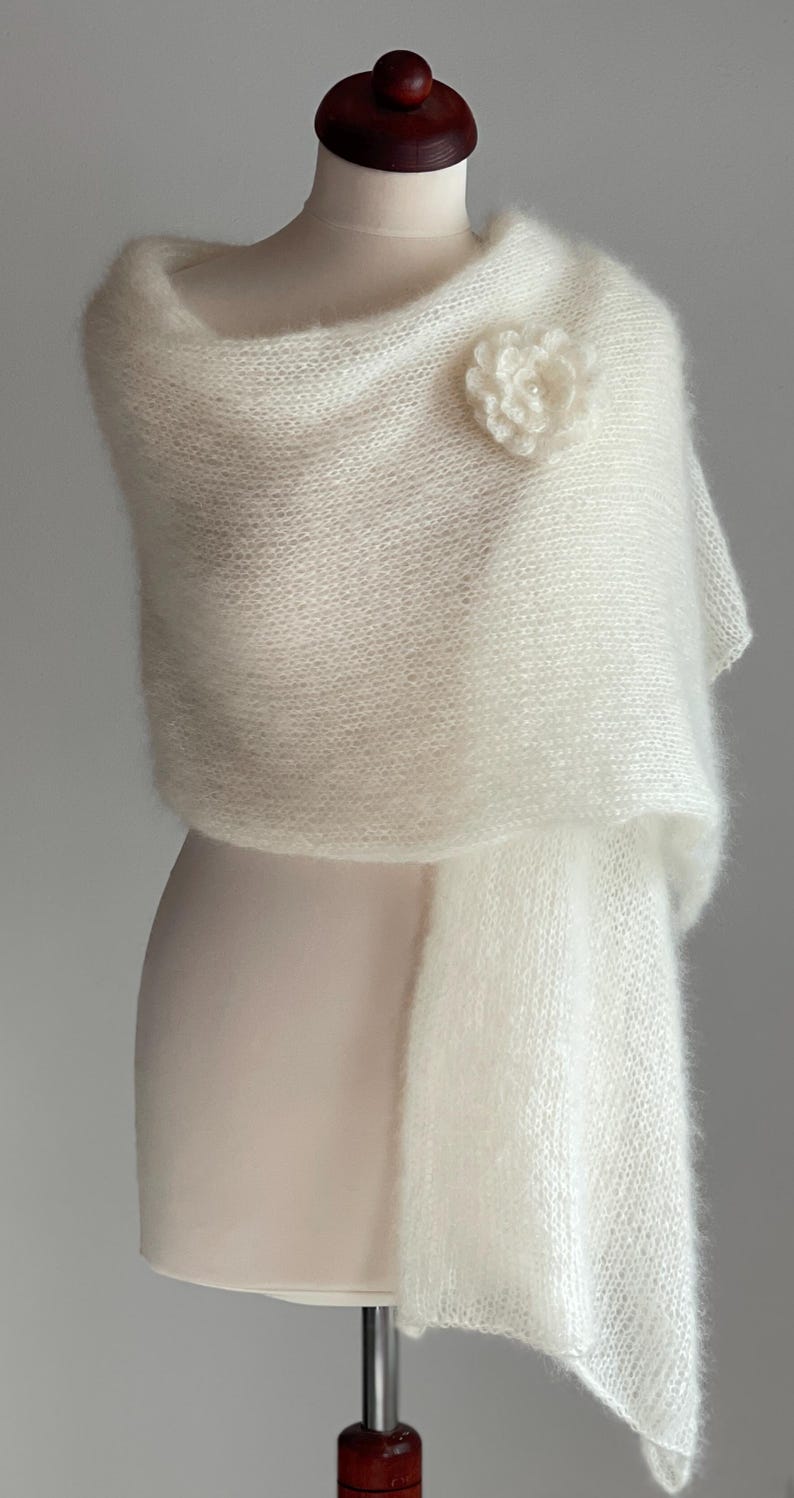 May include: A white, fluffy shawl draped over a mannequin. The shawl is secured with a flower-shaped brooch. The fabric appears soft and delicate, with a slight sheen. The overall aesthetic is elegant and suitable for special occasions.