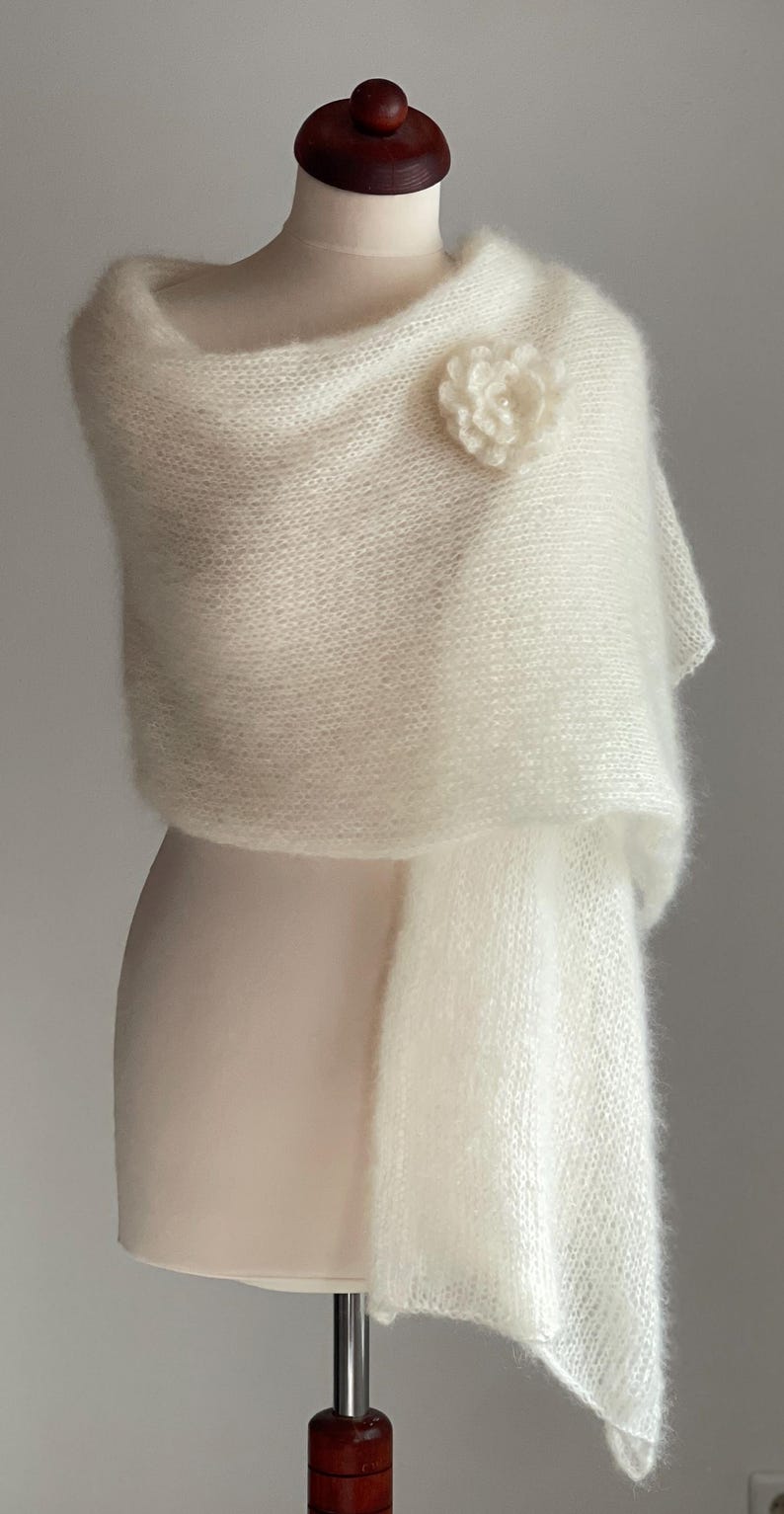 May include: A cream-colored, fluffy shawl draped over a mannequin. The shawl is adorned with a matching flower pin. The fabric appears soft and delicate, with a slight sheen. The overall style is elegant and suitable for special occasions.