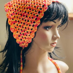 May include: A handmade crochet headscarf in shades of orange, yellow, and pink. The head covering has a scalloped edge and a tie that hangs down the back. The crochet pattern is open, creating a lacy effect. The headscarf is a vibrant accessory.
