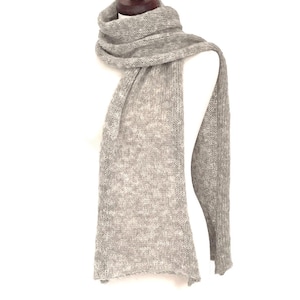 May include: A light gray, knitted scarf draped over a white mannequin. The scarf is made of a soft, textured material and has a long, rectangular shape. The scarf is wrapped around the mannequin's neck and hangs down on both sides.