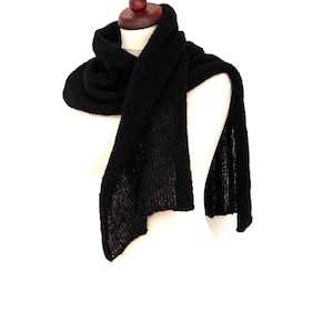 May include: A black knitted scarf draped over a mannequin. The scarf is made of a soft, textured material and is displayed against a white background. The scarf is a solid black color and appears to be warm and cozy.