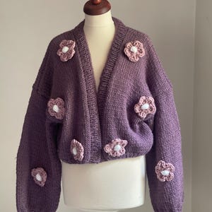 Puede incluir: Un cárdigan morado de punto grueso con cuello en V y mangas largas. El suéter está adornado con múltiples aplicaciones de flores de ganchillo en tonos rosa y blanco. El cárdigan se exhibe en un maniquí.