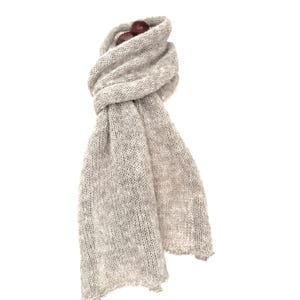 May include: A light gray knitted scarf with a soft, textured appearance. The scarf is draped and knotted, with two small, dark brown beads at the top. The ends of the scarf are visible, and the background is white.