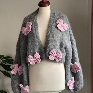 May include: A grey, cropped cardigan with pink bow embellishments. The open-front sweater has long sleeves and is displayed on a mannequin. The bows are evenly spaced across the front and sleeves of the garment.