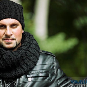 May include: A person wearing a black knit beanie and a black infinity scarf. The person is also wearing a black leather jacket. The background is blurred green foliage.