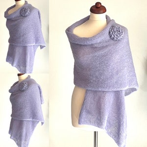 May include: A light purple shawl with a flower accent, displayed on a mannequin. The shawl has a draped design with a cowl neck and a rectangular bottom. The flower is a matching light purple color.