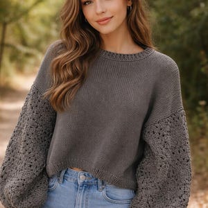 Puede incluir: Un suéter gris con mangas de ganchillo y cuello redondo. Se combina con vaqueros azul claro. Las mangas presentan un diseño calado, añadiendo textura. El estilo es informal y cómodo, ideal para el día a día.