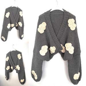 Könnte beinhalten: Eine dunkelgraue Strickjacke mit V-Ausschnitt und langen Ärmeln. Das Kleidungsstück ist mit mehreren cremefarbenen, strukturierten, wolkenförmigen Applikationen verziert, die über die Vorderseite und die Ärmel verteilt sind. Der Strick hat eine Rippstruktur.