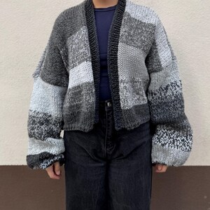 Könnte beinhalten: Ein grauer und weißer Strickcardigan mit langen Ärmeln und lockerer Passform. Der Cardigan hat ein Patchwork-Design mit verschiedenen Grautönen und Weiß.