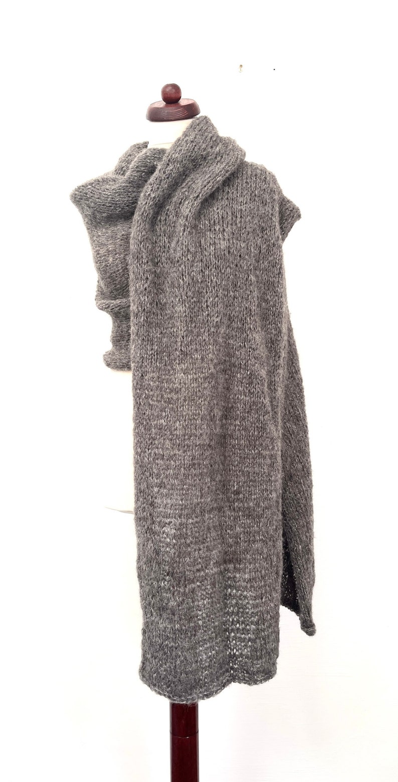 May include: A gray knitted scarf displayed on a mannequin. The scarf is a solid color, with a thick, textured knit. The scarf is long and drapes down the front of the mannequin. The scarf is made of a soft, warm material.
