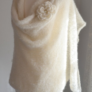 May include: A white knitted shawl with a delicate crocheted flower embellishment. The shawl is draped over a white mannequin and has a soft, flowing texture.