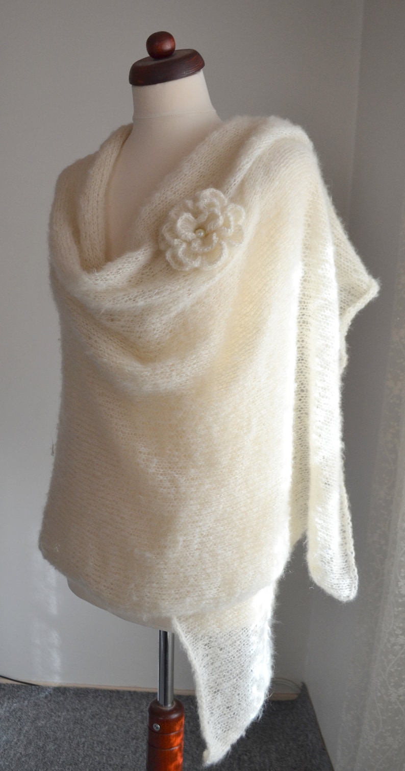 May include: A white knitted shawl with a delicate crocheted flower embellishment. The shawl is draped over a white mannequin and has a soft, flowing texture.