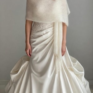 May include: A cream-colored bridal gown with a satin skirt and a lace bodice. The bride is wearing a fluffy, off-white shawl and a flower pin. The dress has a flowing design, perfect for a wedding or special occasion.