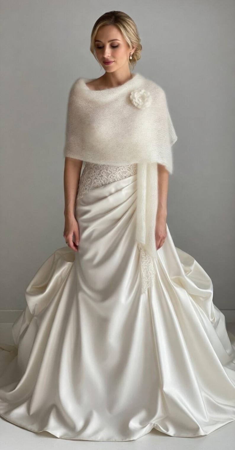 May include: A cream-colored bridal gown with a satin skirt and a lace bodice. The bride is wearing a fluffy, off-white shawl and a flower pin. The dress has a flowing design, perfect for a wedding or special occasion.