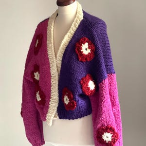 May include: A handmade, chunky knit cardigan in a color-blocked design. The sweater features a purple and magenta body with cream-colored trim and red and white flower appliqués. The sleeves are magenta. The cardigan is displayed on a mannequin.