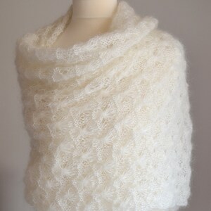 May include: Ivory-colored, open-weave shawl draped over a mannequin. The shawl has a delicate, textured pattern and is made of a soft, fluffy material. The shawl is displayed on a mannequin with a wooden top.